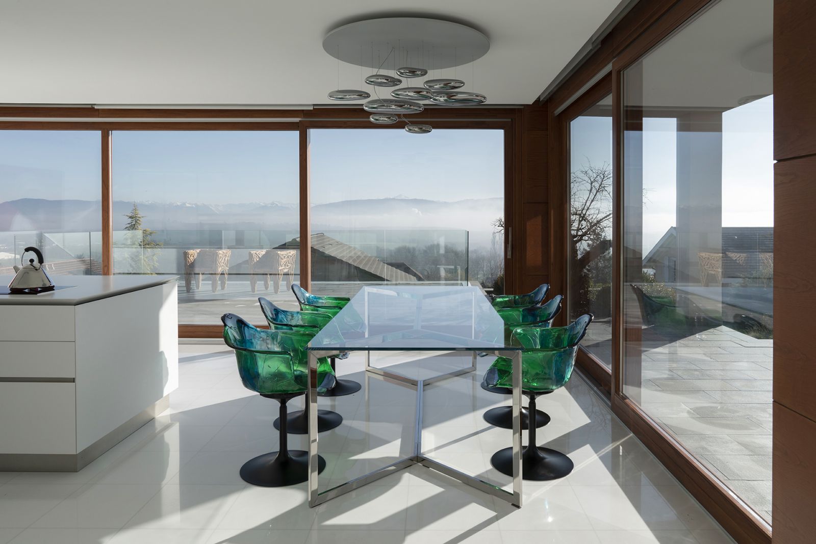 Stuhl «Ella» in Grün- und Blautönen bringt Farbe in den Küchenbereich, ohne die Transparenz des Hauses zu stören. Design: Jacopo Foggini für Edra. Leuchte «Mercury» von Ross Lovegrove für Artemide. Auf der Terrasse sind die Stühle «Favela» aus naturbelassenen Holzleisten zu sehen. Designer: Fernando und Humberto Campana für Edra.