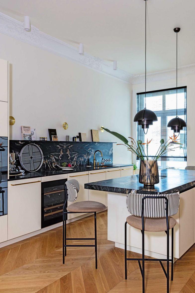 Die Accessoires aus Messing und die gestreiften Bezüge der Barhocker erinnern an die Zeit des Art déco. Dazu kombinieren die Innenarchitektinnen bewegte Oberflächen aus dunklem Naturstein, cremefarbene Fronten und schwarze Pendelleuchten von Gubi.